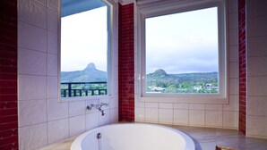 A bathroom with a large bathtub, two windows with views of a mountainous landscape, and red-tiled walls. - Millennium Inn (Hengchun)
