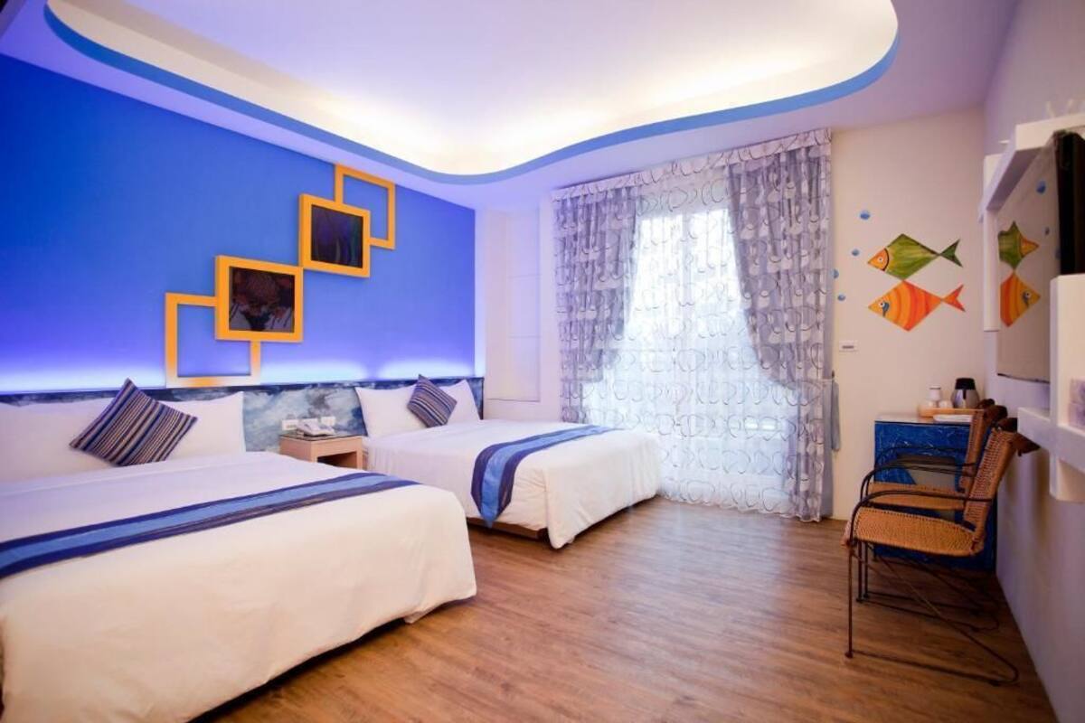 Habitació d'hotel amb dos llits, un terra de fusta, un escriptori amb cadira i una finestra amb cortines estampades.