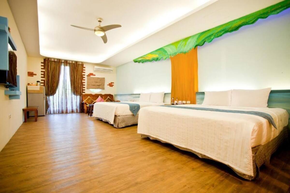 Habitació d'hotel amb dos llits, un ventilador de sostre, una taula de treball amb cadira i una finestra amb cortines.