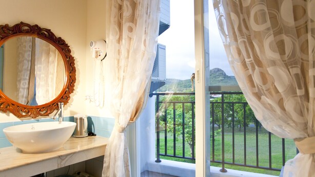A bathroom with a large oval mirror, a white sink, and a hairdryer on the counter. There is a balcony with a view of greenery and a building in the background. - Millennium Inn (Hengchun)