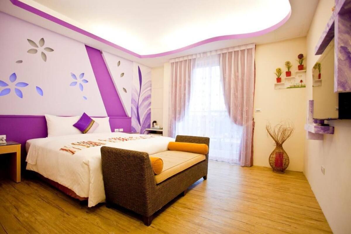 Un dormitori amb un llit, un sofà, una tauleta de nit, un televisor fixat a la paret, una finestra amb cortines i una paret amb prestatgeries de flors.