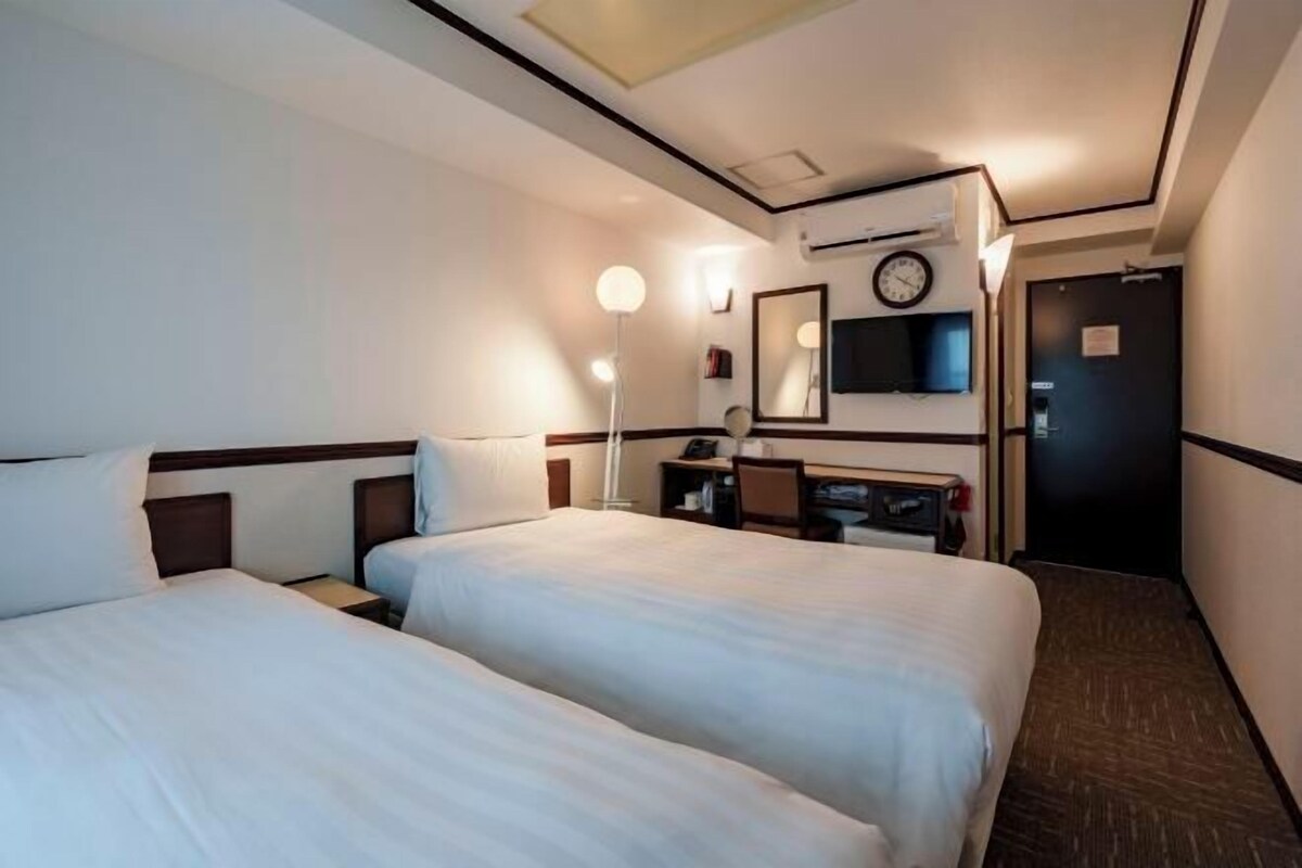 A hotel room with two beds, a desk, a chair, a lamp, a clock, and a door.