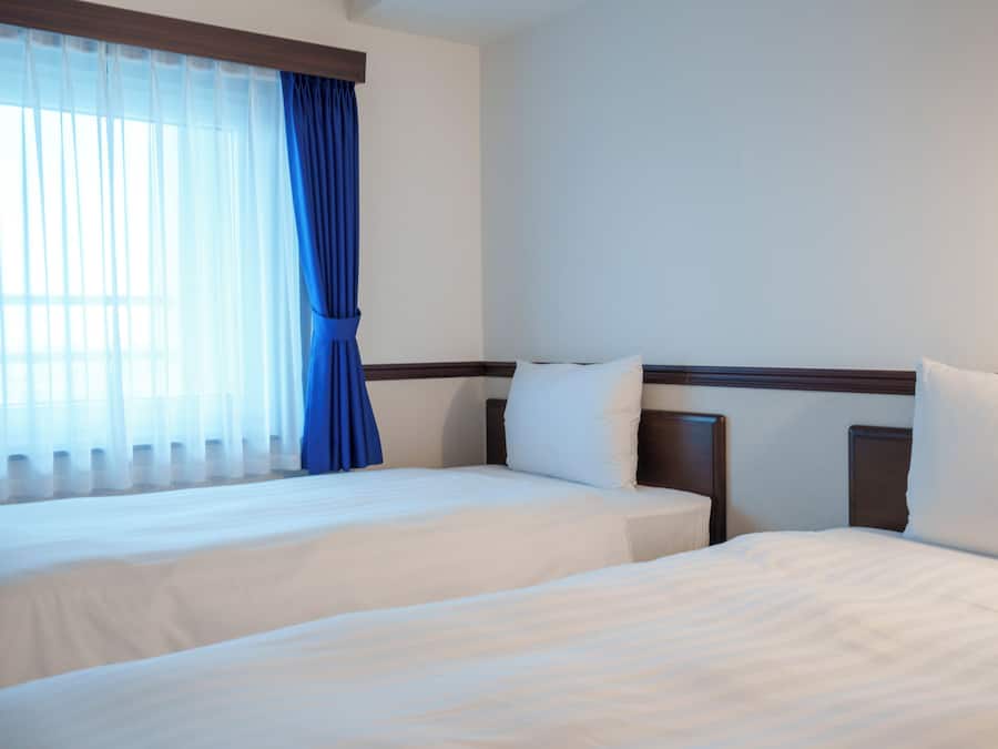 A hotel room with two beds, a large window with sheer curtains, and a plain white wall.