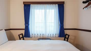 Quarto de hotel com cama, duas cadeiras, uma mesinha e janela com cortinas.