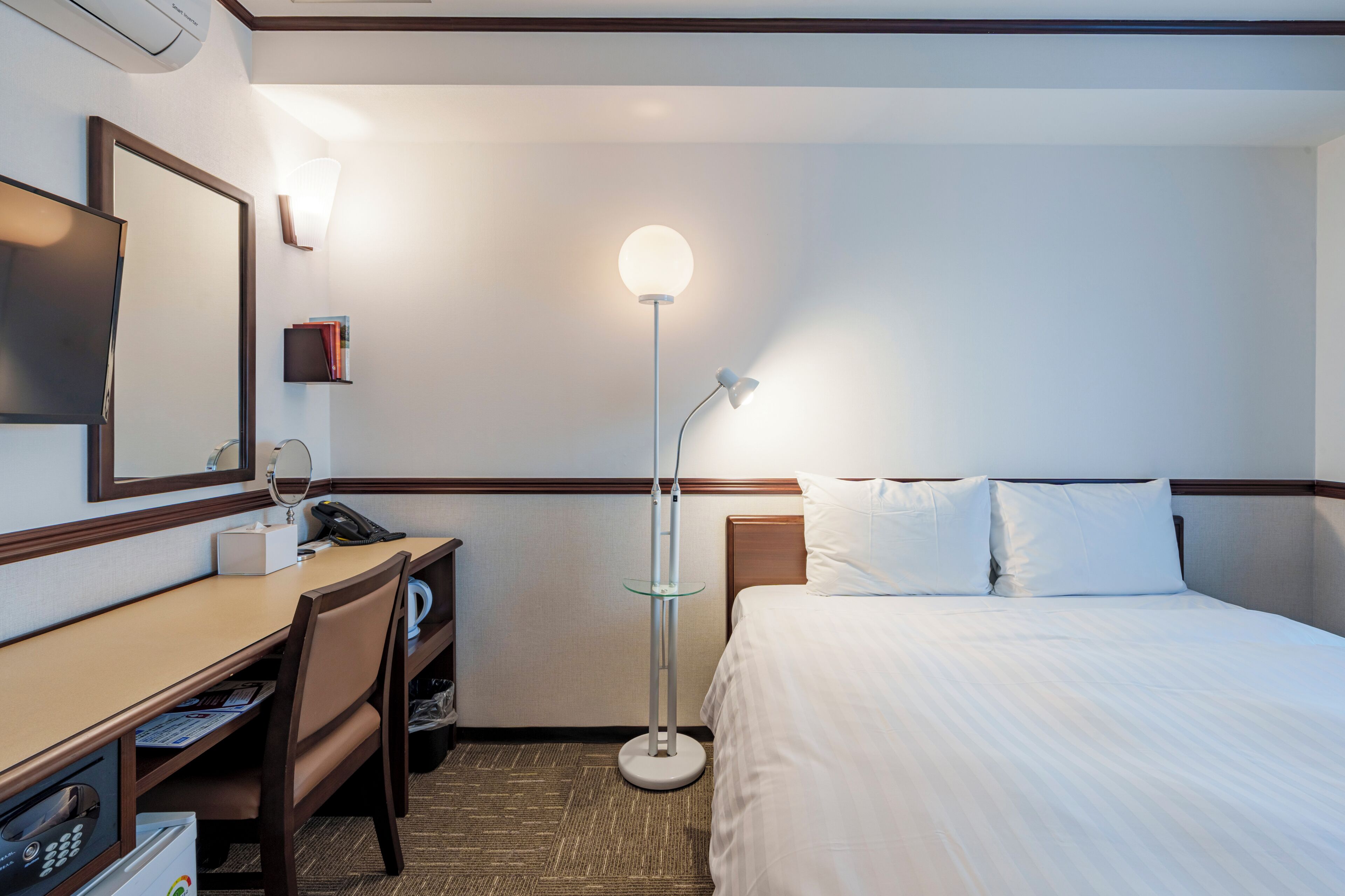 A hotel room with a bed, desk, chair, and a standing lamp.