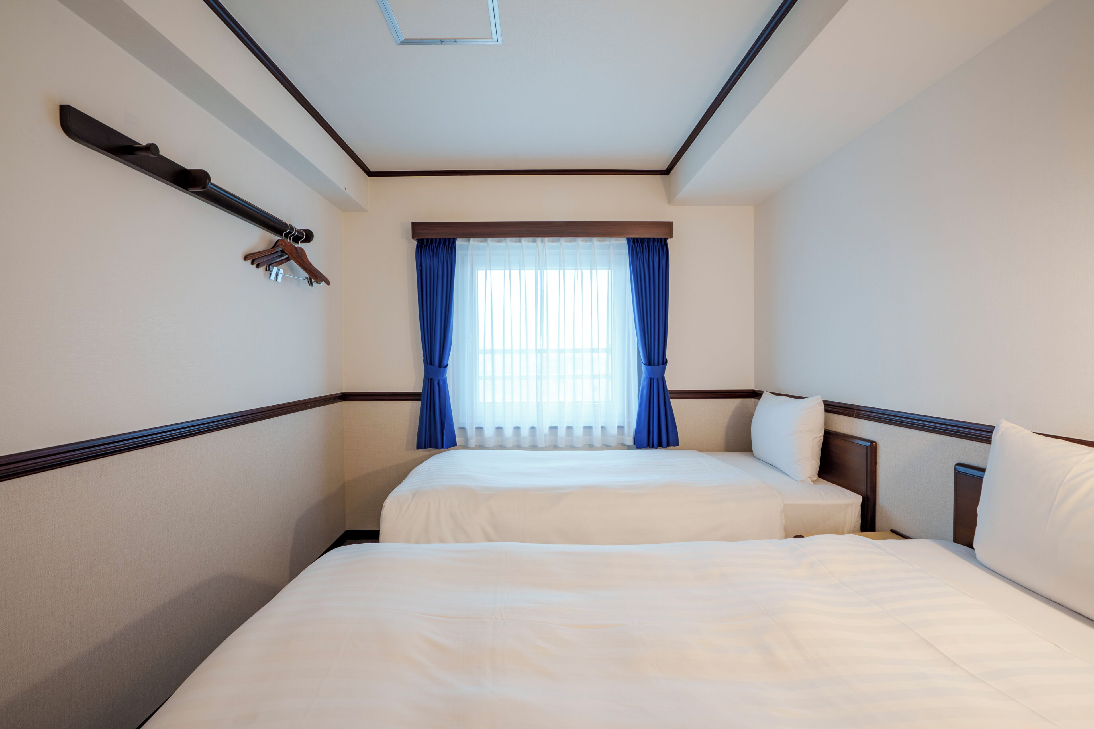 A hotel room with two beds, a window with blue curtains, a coat rack, and a ceiling light.