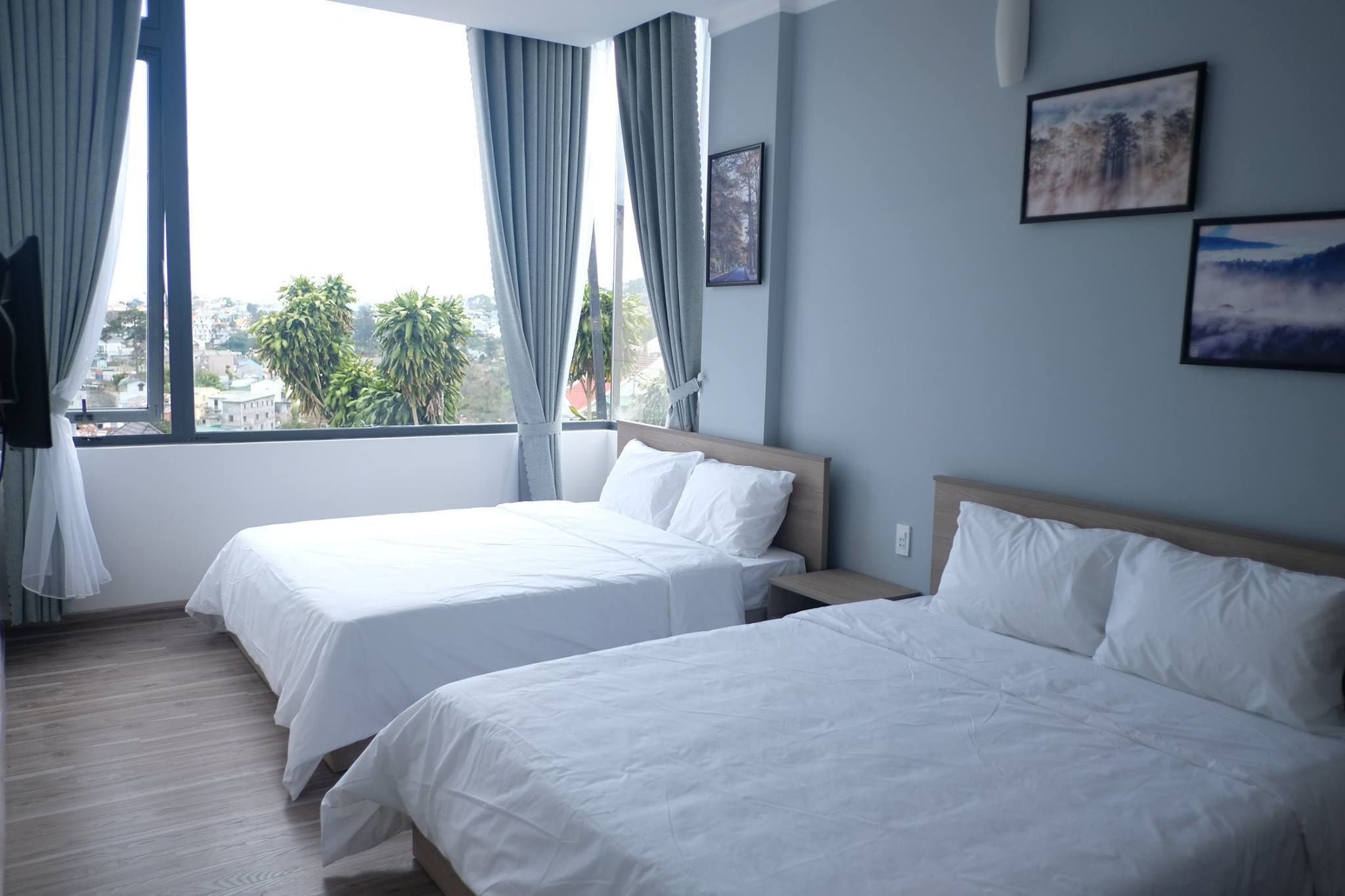 A hotel room with two beds, a large window with curtains, and two framed pictures on the wall.