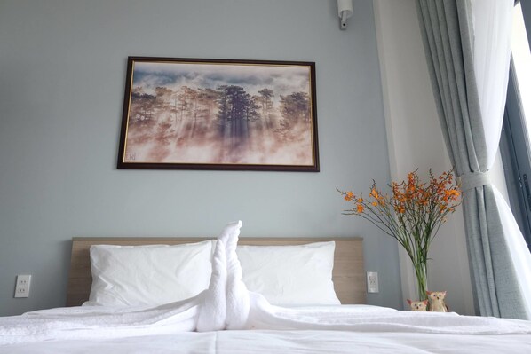 A neatly made bed with white linens, a framed painting of a forest scene, and a vase with orange flowers on a bedside table. - Dalat Memory Inn (Xuan Huong - Da Lat)