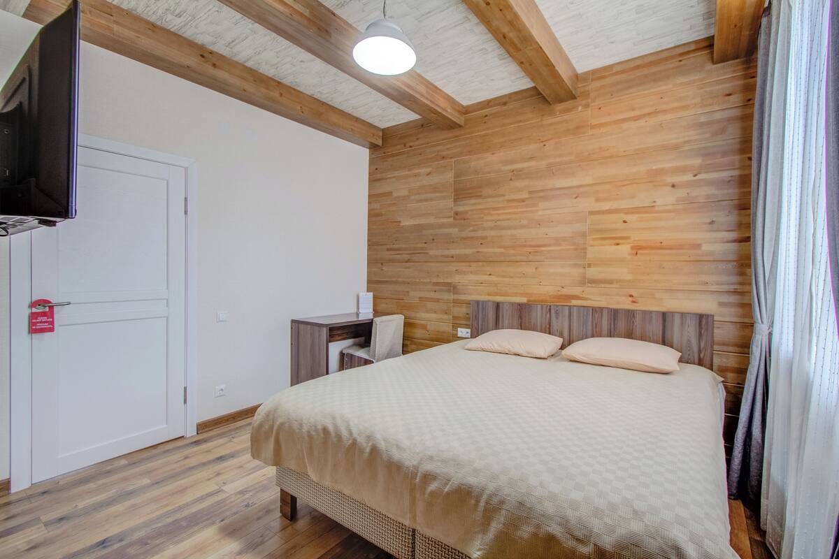 A bedroom with a wooden headboard, a bed with a beige bedspread, a desk with a chair, a television mounted on the wall, and a white door.