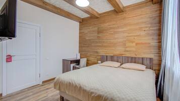 Un dormitorio con cabecera de madera, cama con colcha color beige, escritorio con silla, televisor montado en la pared y una puerta blanca.