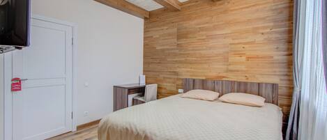 Un dormitorio con cabecera de madera, cama con colcha color beige, escritorio con silla, televisor montado en la pared y una puerta blanca.