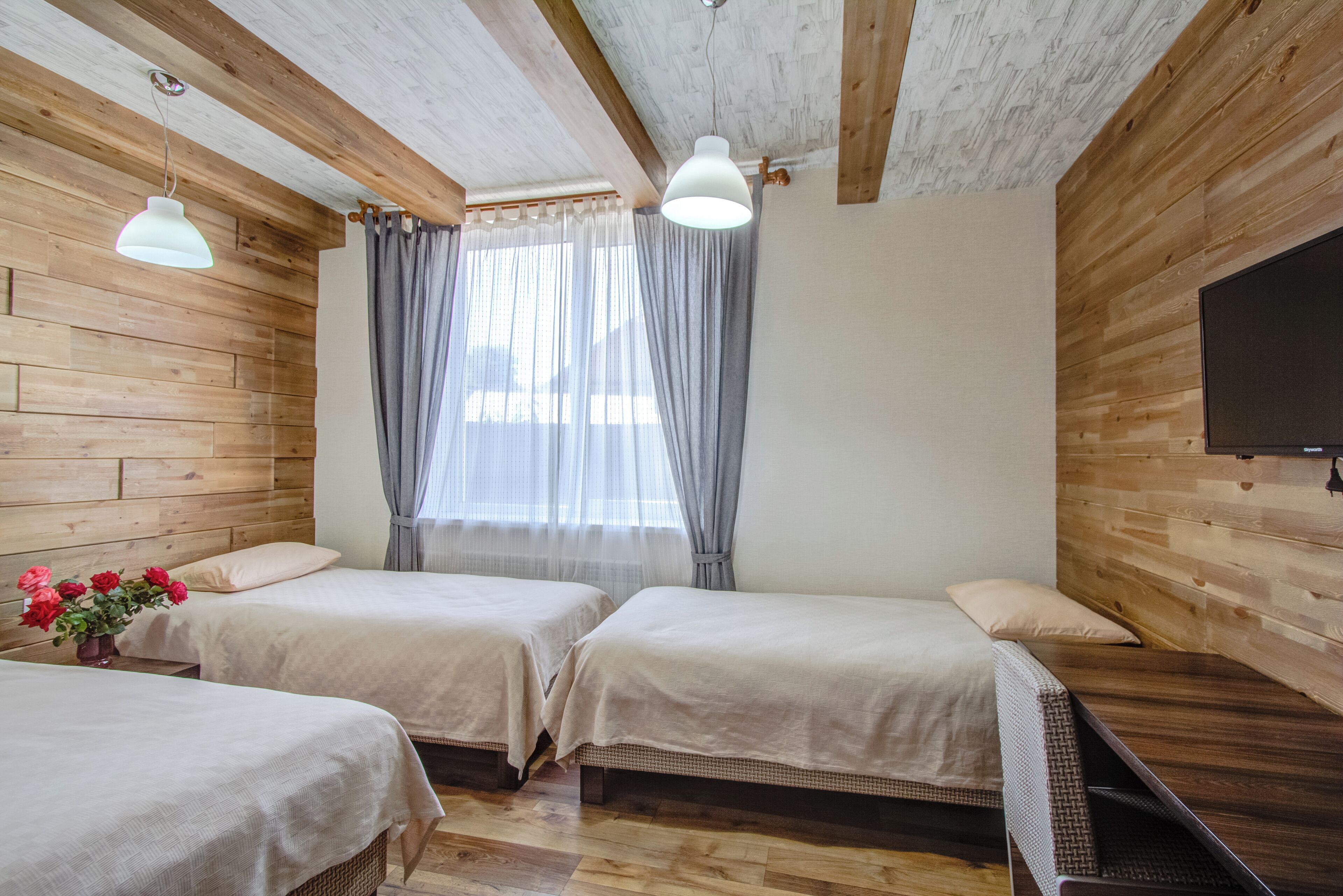 Habitación con dos camas, paredes de madera, techo con vigas a la vista, un ventanal con cortinas y un televisor de pantalla plana montado en la pared.