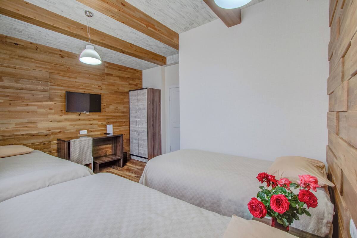 A bedroom with two beds, a wooden ceiling, a flat-screen TV, a desk, and a vase with flowers.