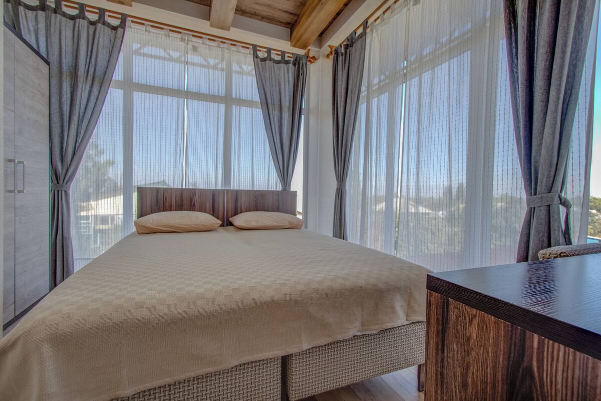 A bedroom with a large bed, wooden headboard, and a view through sheer curtains.