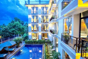 Un hotel de varios pisos con piscina en la azotea, balcones y abundante vegetación.