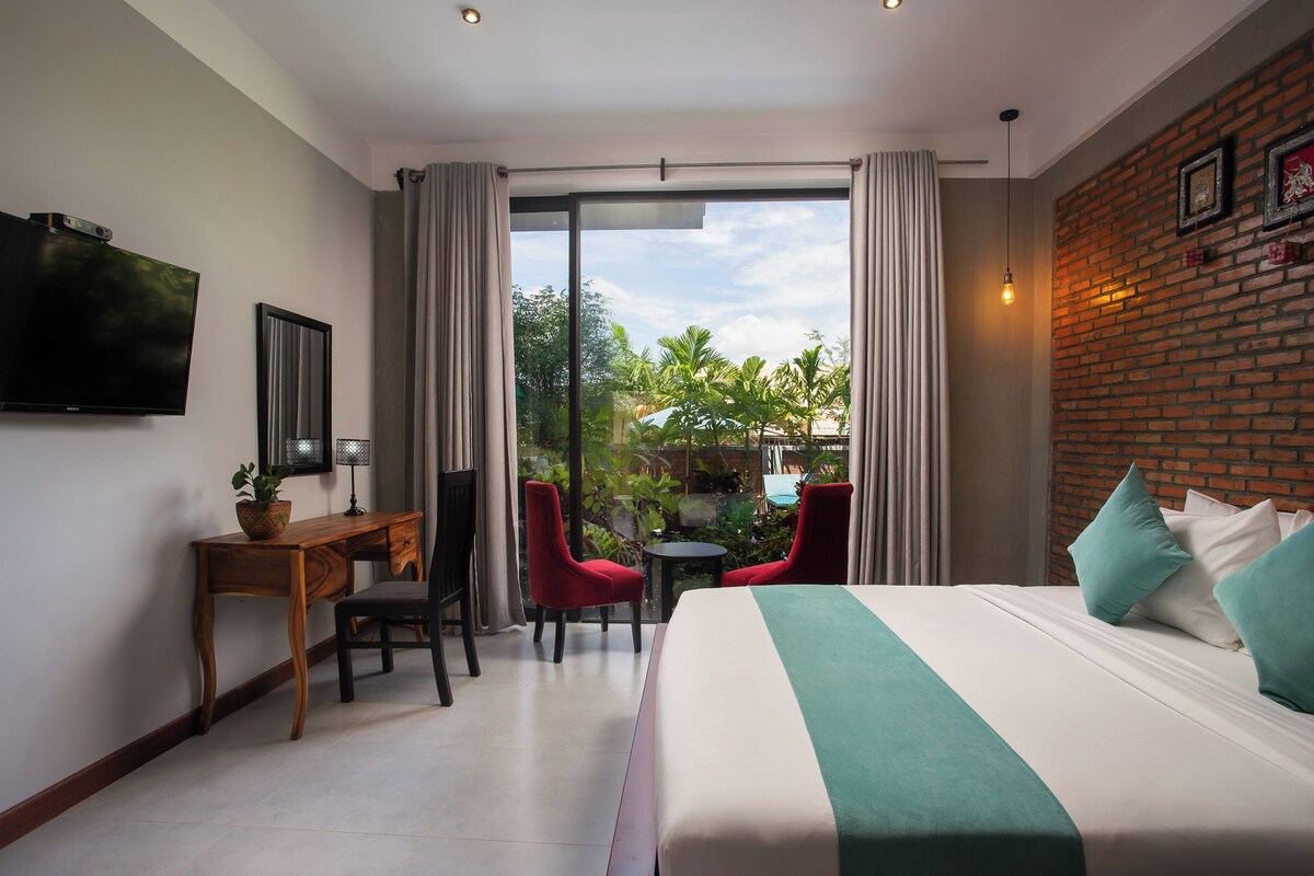 A hotel room with a bed, a desk, two chairs, a TV, and a view of greenery outside.