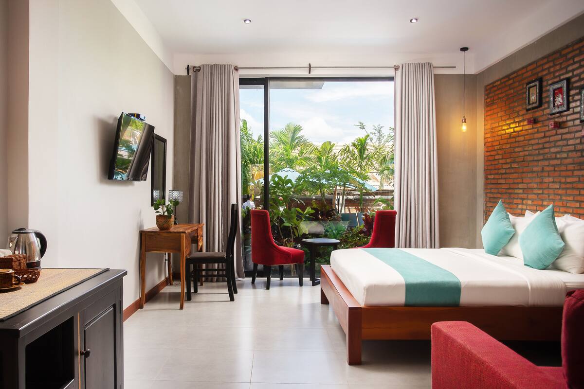 A hotel room with a bed, a desk, a chair, a TV, and a view of greenery outside.