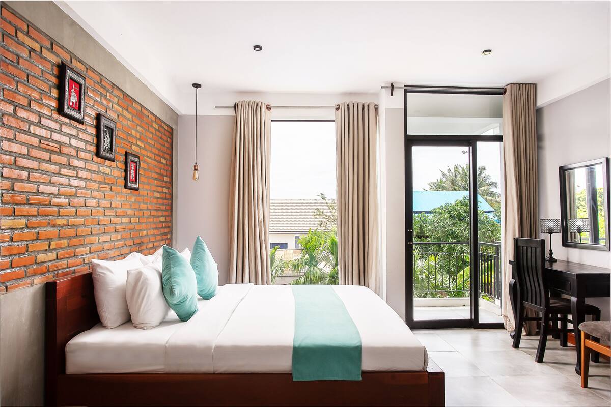 A modern hotel room with a brick accent wall, a large bed with white bedding and a teal throw, a desk with a chair, and a balcony with a view of greenery.