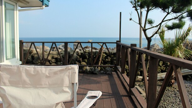 A wooden deck with a white chair and a small table, overlooking a rocky shoreline and the sea. - Sunny Namwon (Seogwipo)