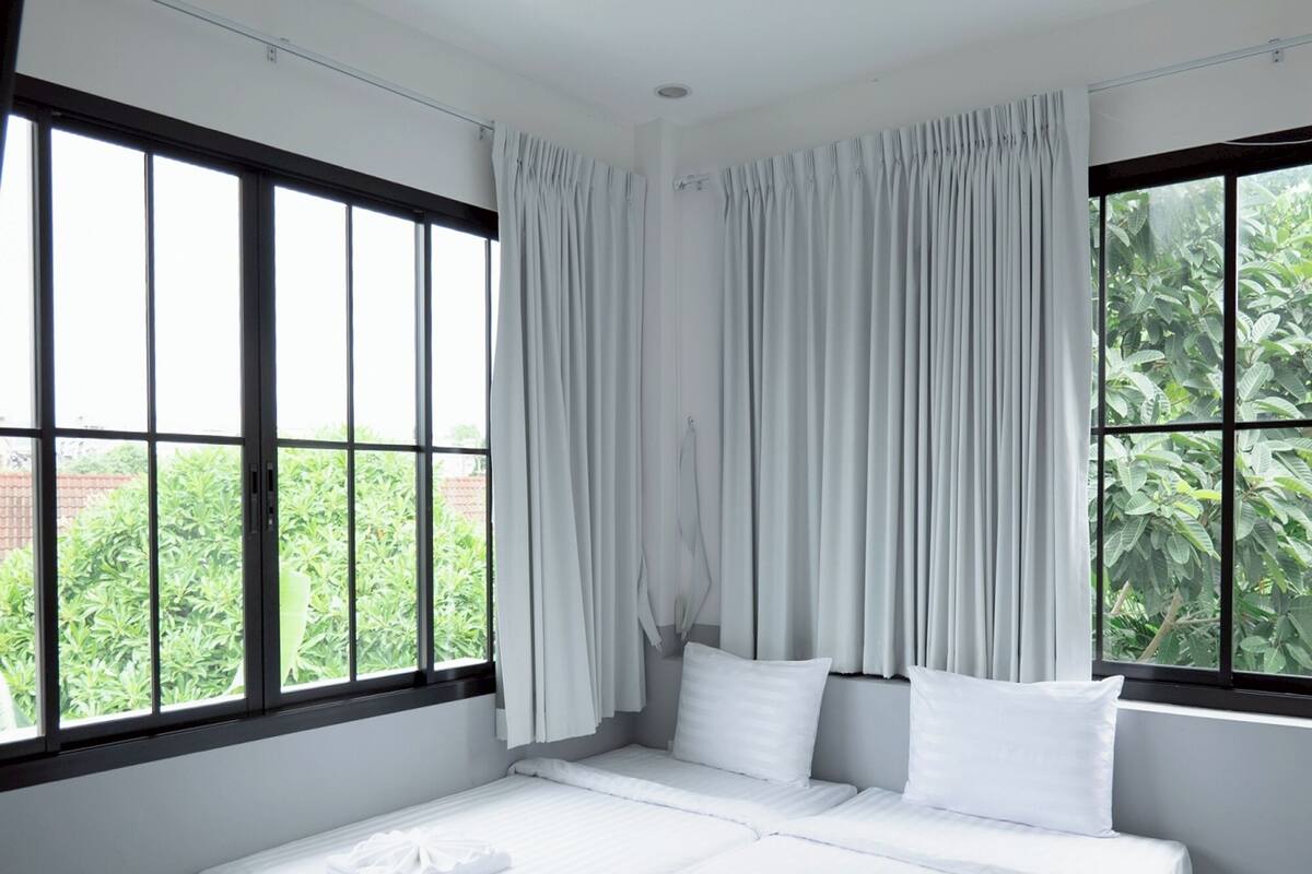 A bedroom with a large bed, two pillows, white curtains, and a view of greenery outside.