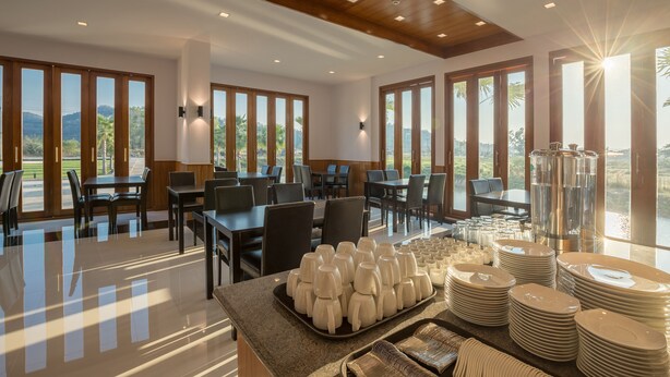Un grand espace dédié à la salle à manger, avec des tables et des chaises, de grandes fenêtres et un buffet où sont disposés assiettes et verres.