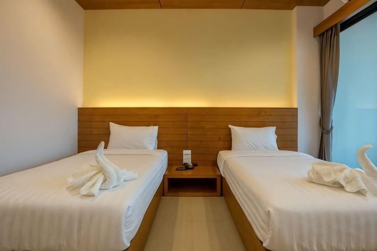 Une chambre d’hôtel avec deux lits simples, une tête de lit en bois, du linge de lit blanc et une table de chevet avec une lampe.