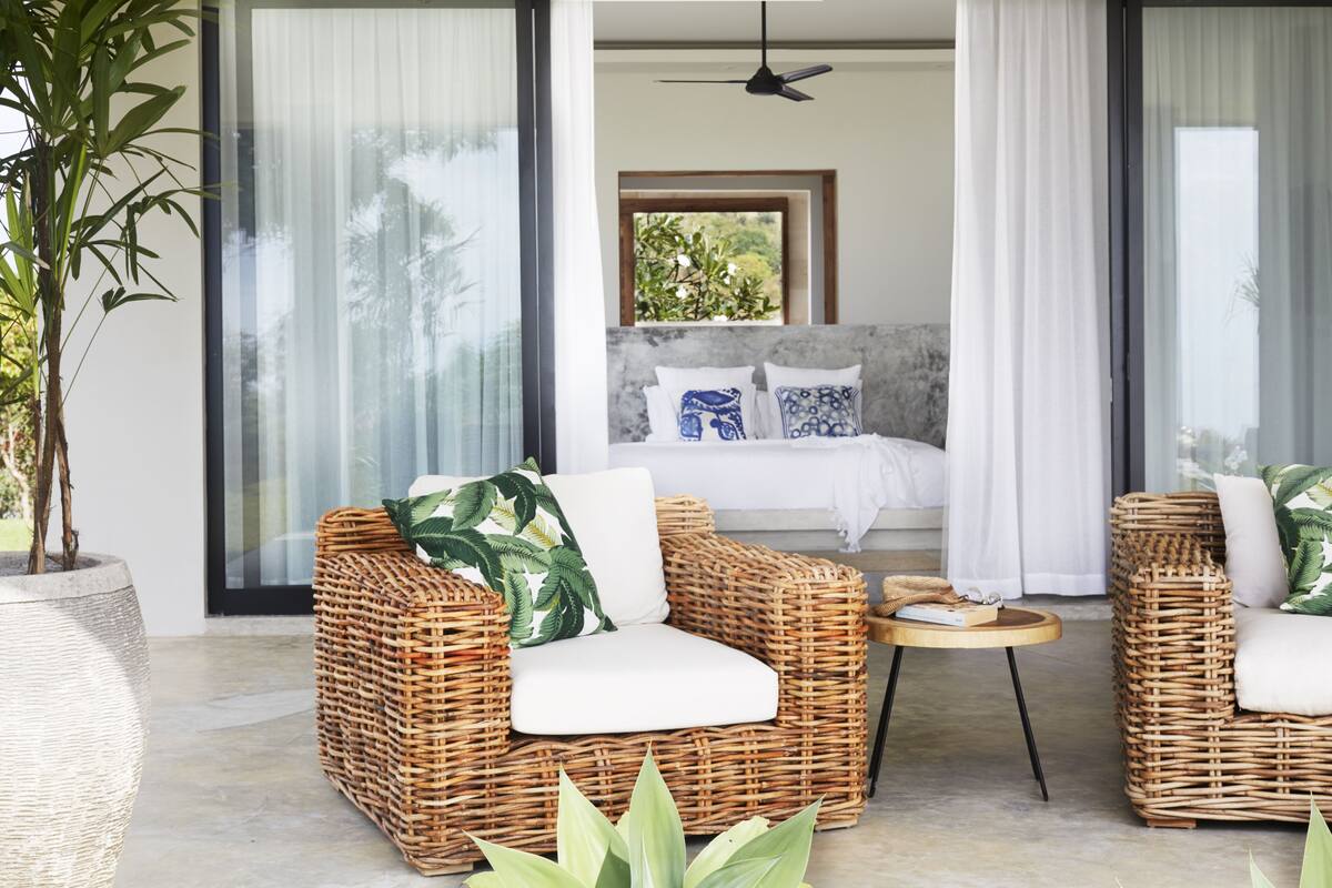 A modern room with wicker furniture, a bed with patterned pillows, and a view of greenery through large windows.