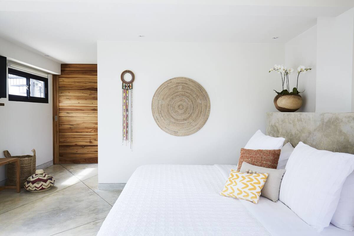 A modern bedroom with a large bed, a wooden door, a wall-mounted woven basket, and a potted plant with white flowers.