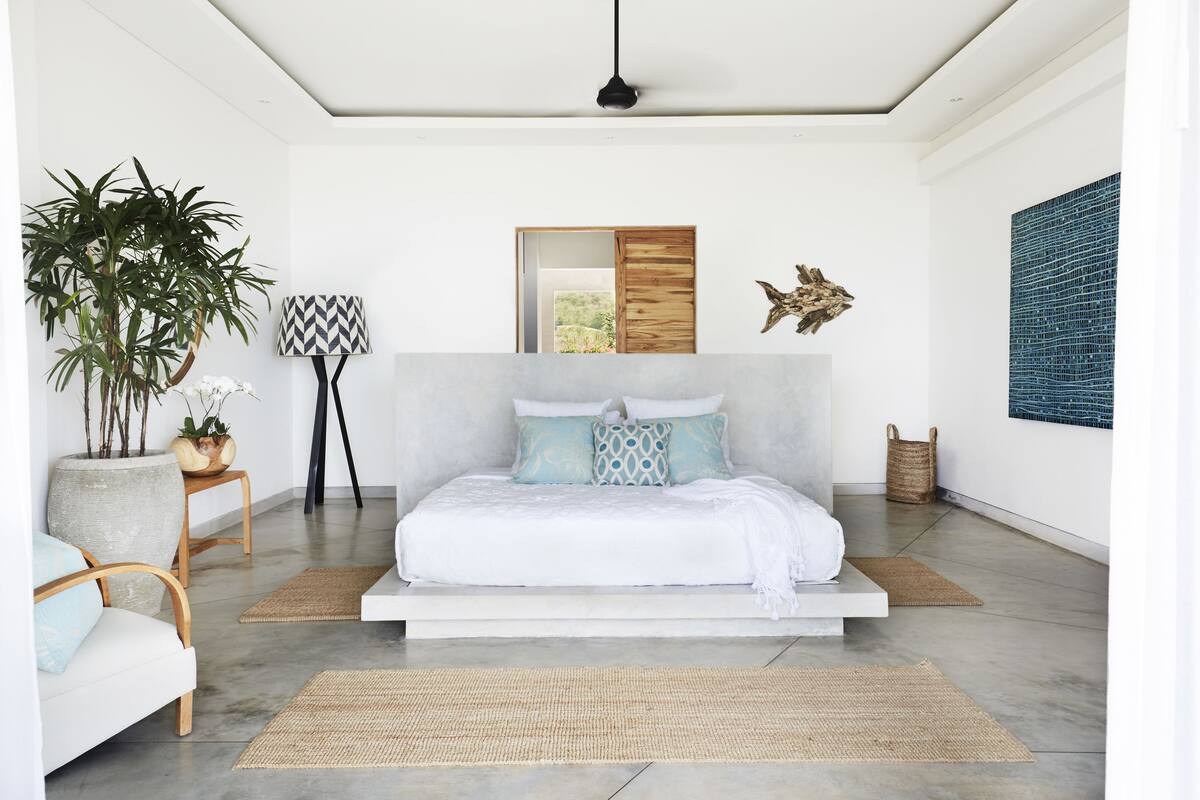 A modern bedroom with a minimalist design, featuring a bed with a wooden headboard, a large potted plant, a small side table, and a woven rug.