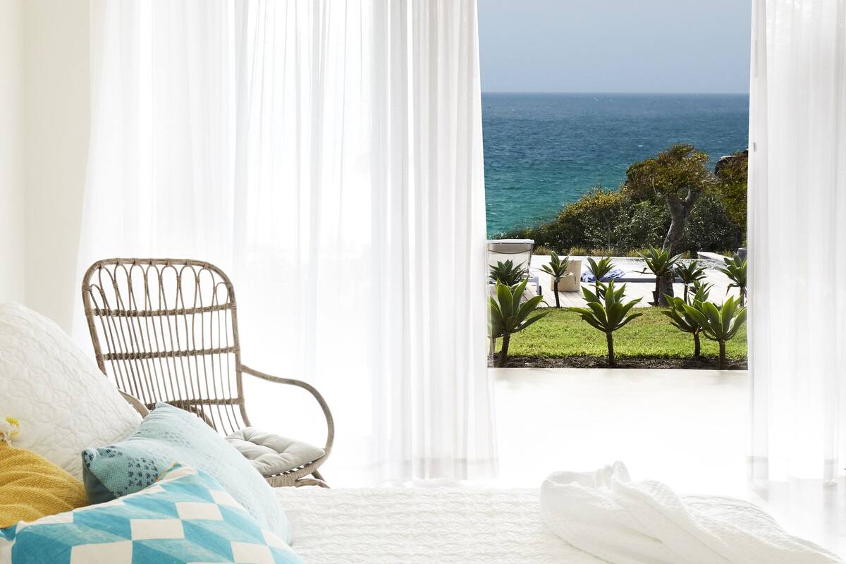 A bedroom with a bed, white curtains, and a view of the ocean.
