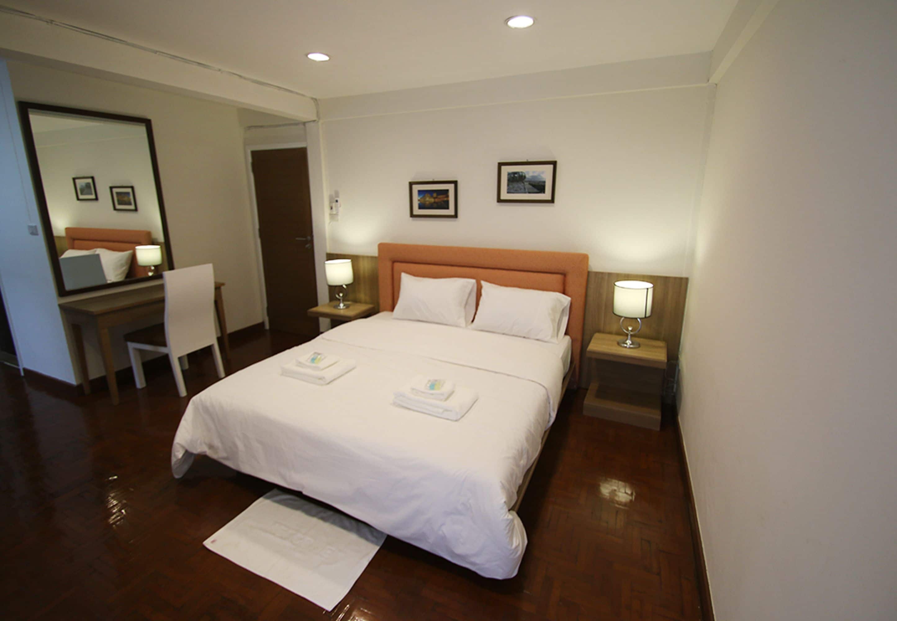 A hotel room with a large bed, two bedside tables with lamps, a desk, and a chair. There are two framed pictures on the wall.