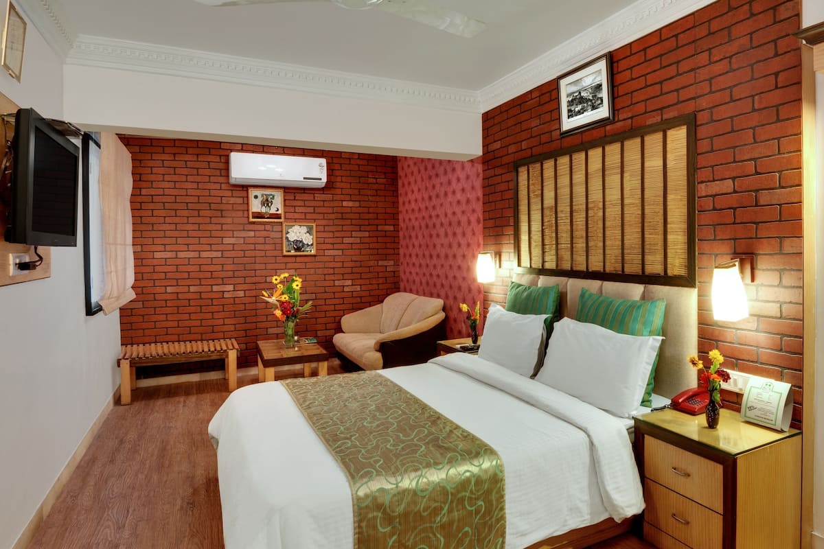 Habitación de hotel con pared de ladrillo, cama con ropa blanca, una silla, una mesita con un florero y una mesita de noche con lámpara y teléfono.