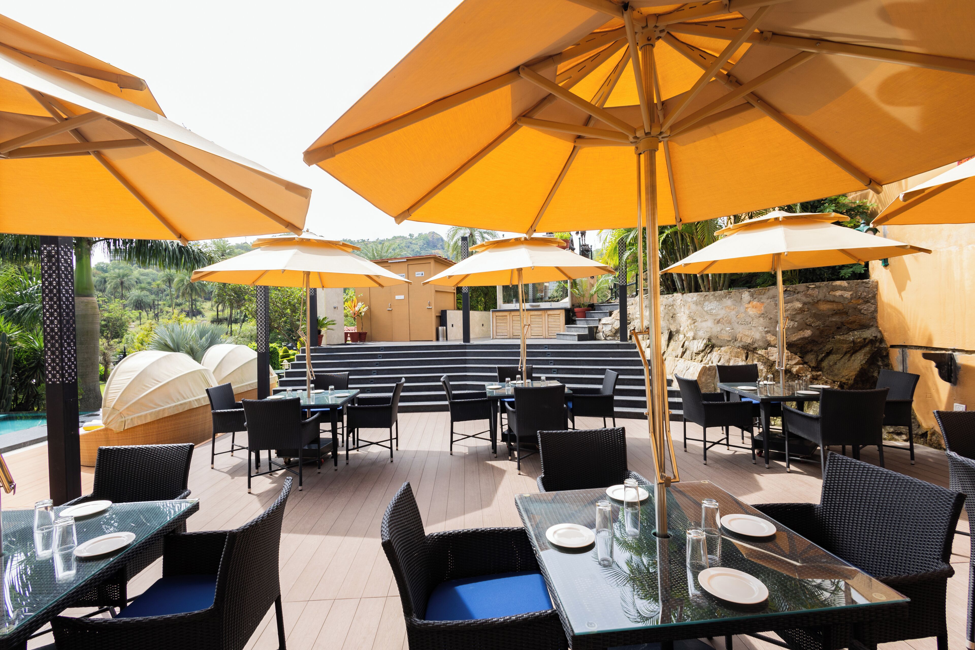 An outdoor dining area with tables and chairs, yellow umbrellas, and a view of a pool and greenery.