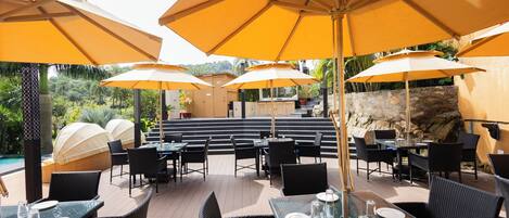 An outdoor dining area with tables and chairs, yellow umbrellas, and a view of a pool and greenery.