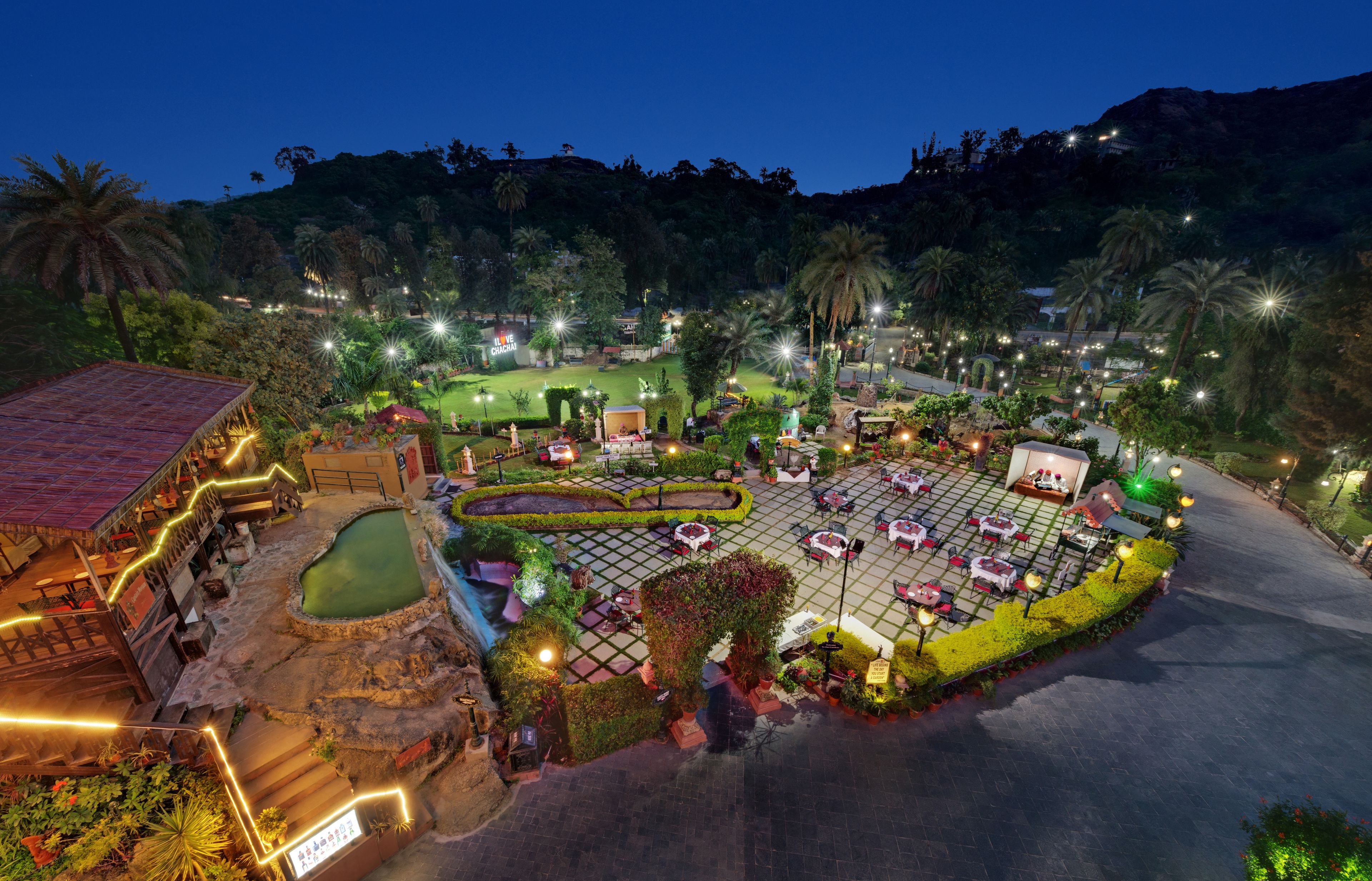 A well-lit outdoor area with a pool, seating, and a variety of plants and trees.