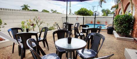 An outdoor patio with round tables and chairs, a pool in the background, and a brick wall.