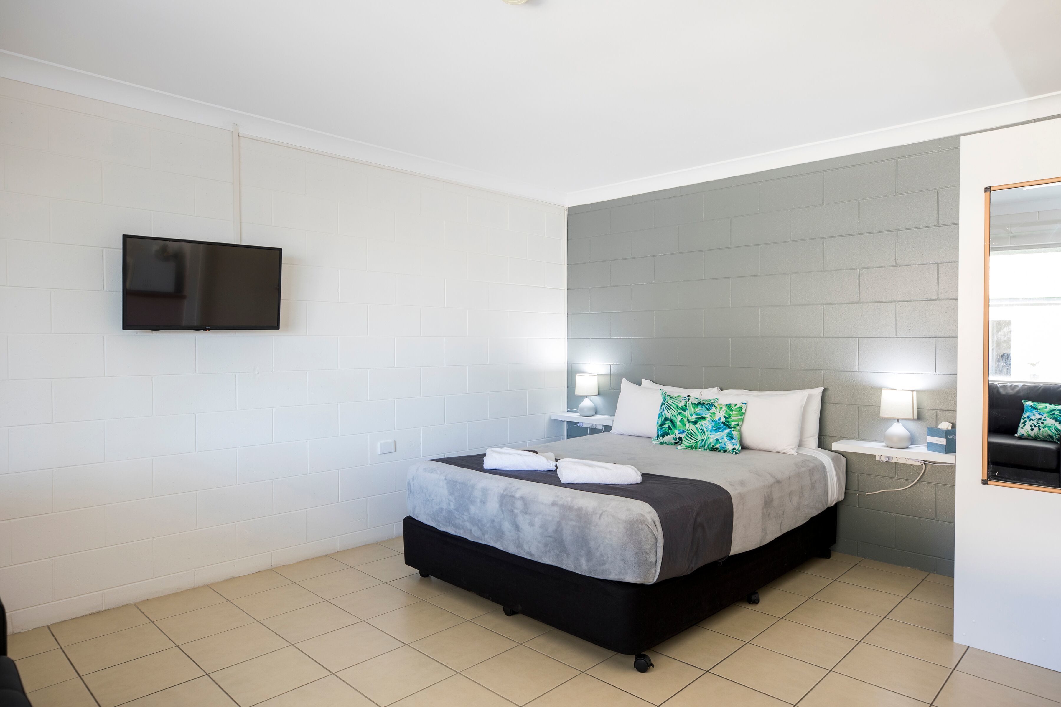 Una habitación de hotel con una cama, un televisor de pantalla plana montado en la pared, una mesita de noche con una lámpara y una ventana con vistas.