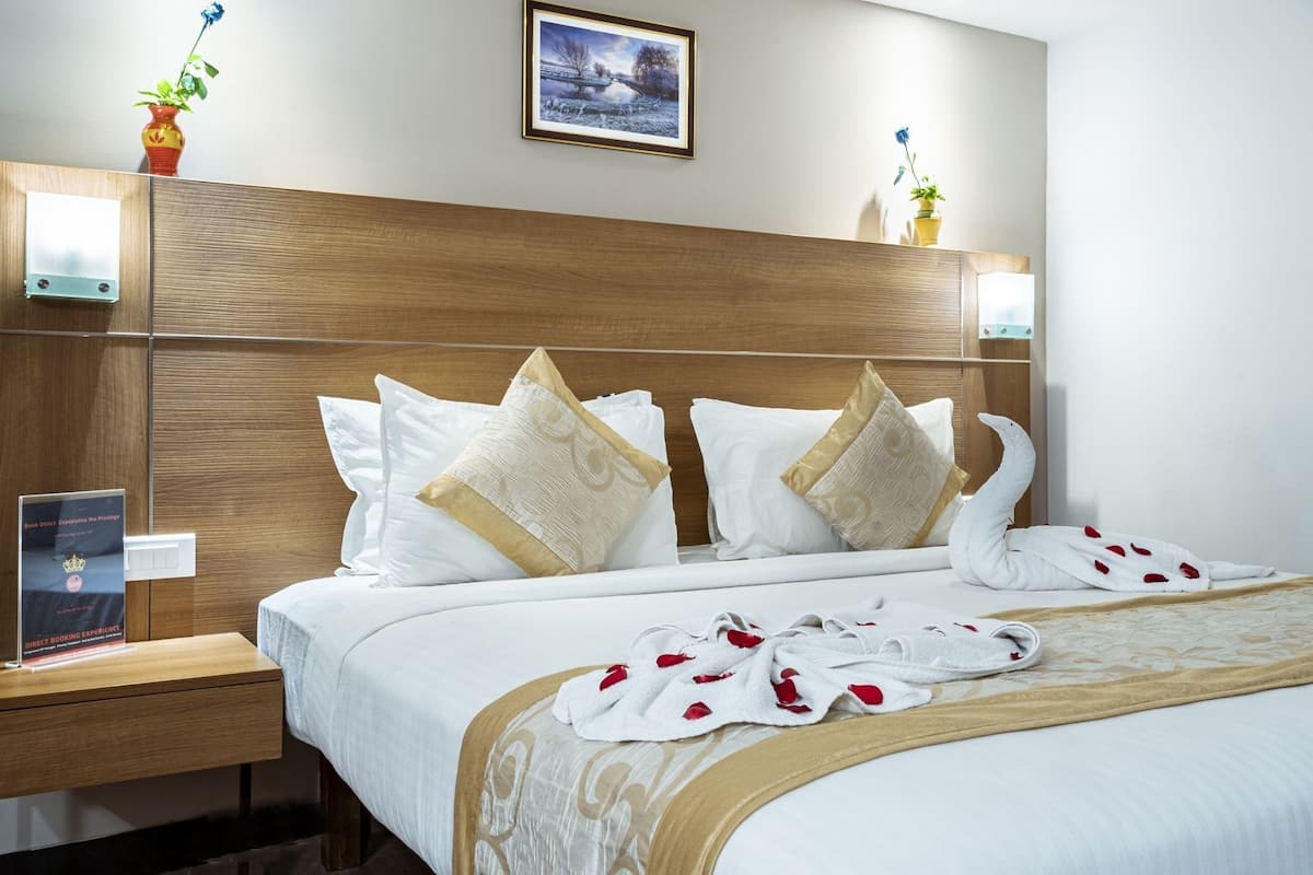 A hotel room with a wooden headboard, a bed with white bedding and decorative pillows, a bedside table with a book, and a framed picture on the wall.