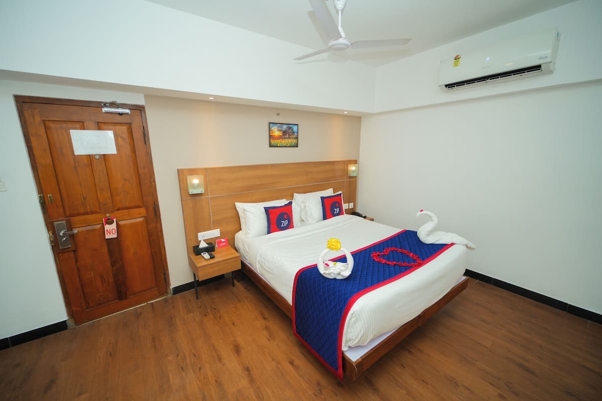 A hotel room with a wooden door, a bed with a blue and red bedspread, a swan decoration, and an air conditioning unit on the wall.