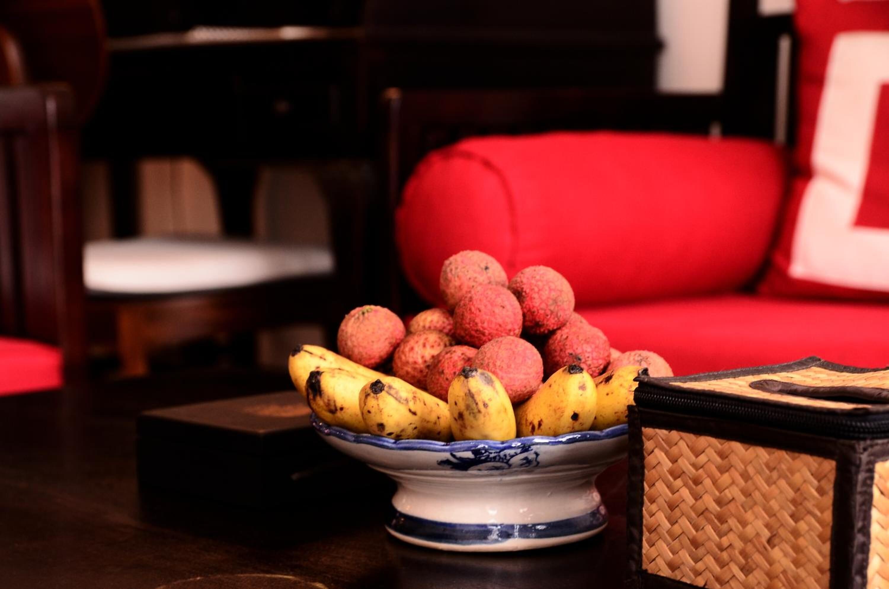 A dining table with a bowl of lychees and bananas, a woven basket, and a red upholstered chair.