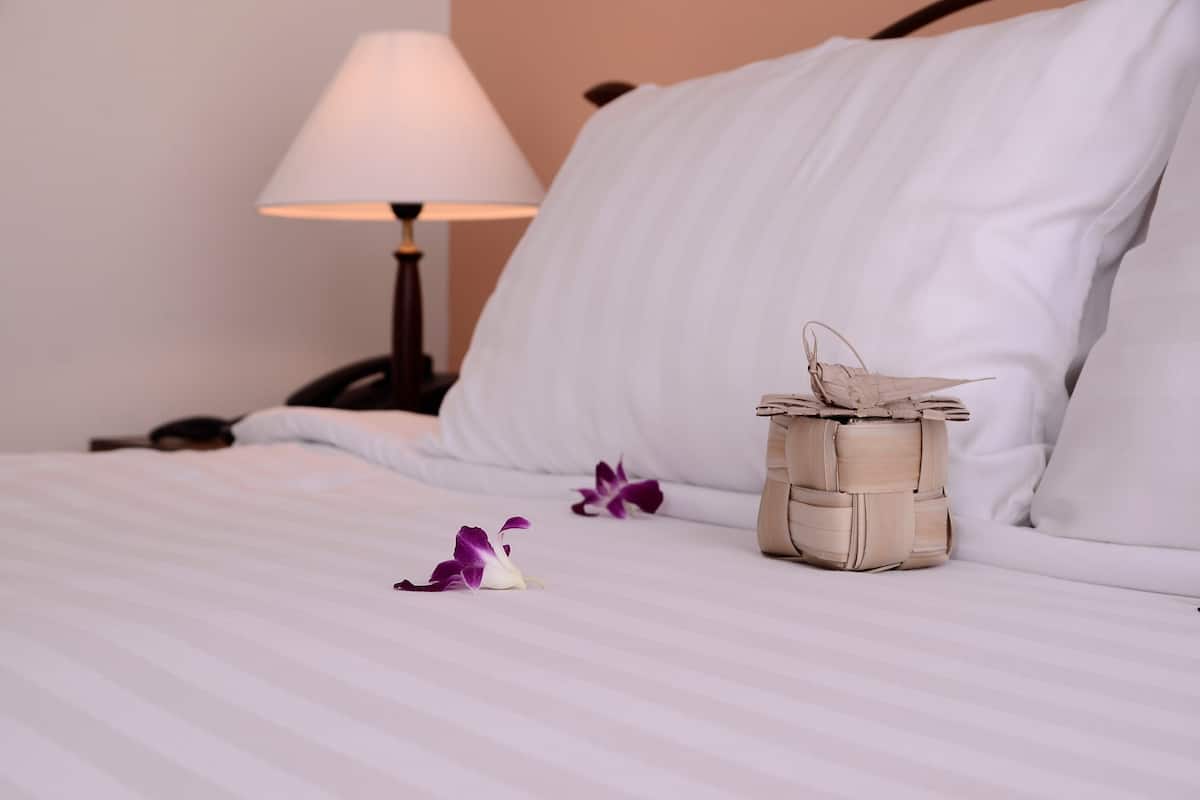 A neatly made bed with white linens, a small woven basket, and a purple flower on the bed.
