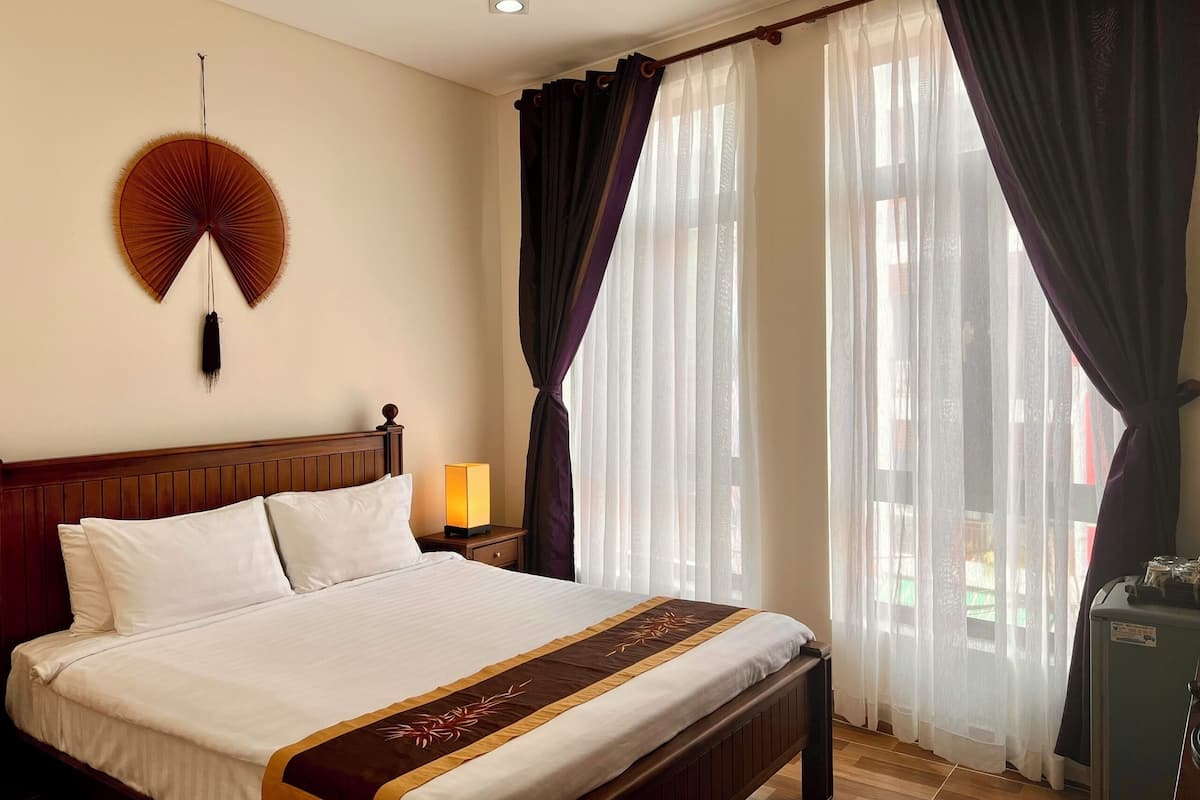 A hotel room with a large bed, a bedside table with a lamp, a wall-mounted fan, and a window with curtains.