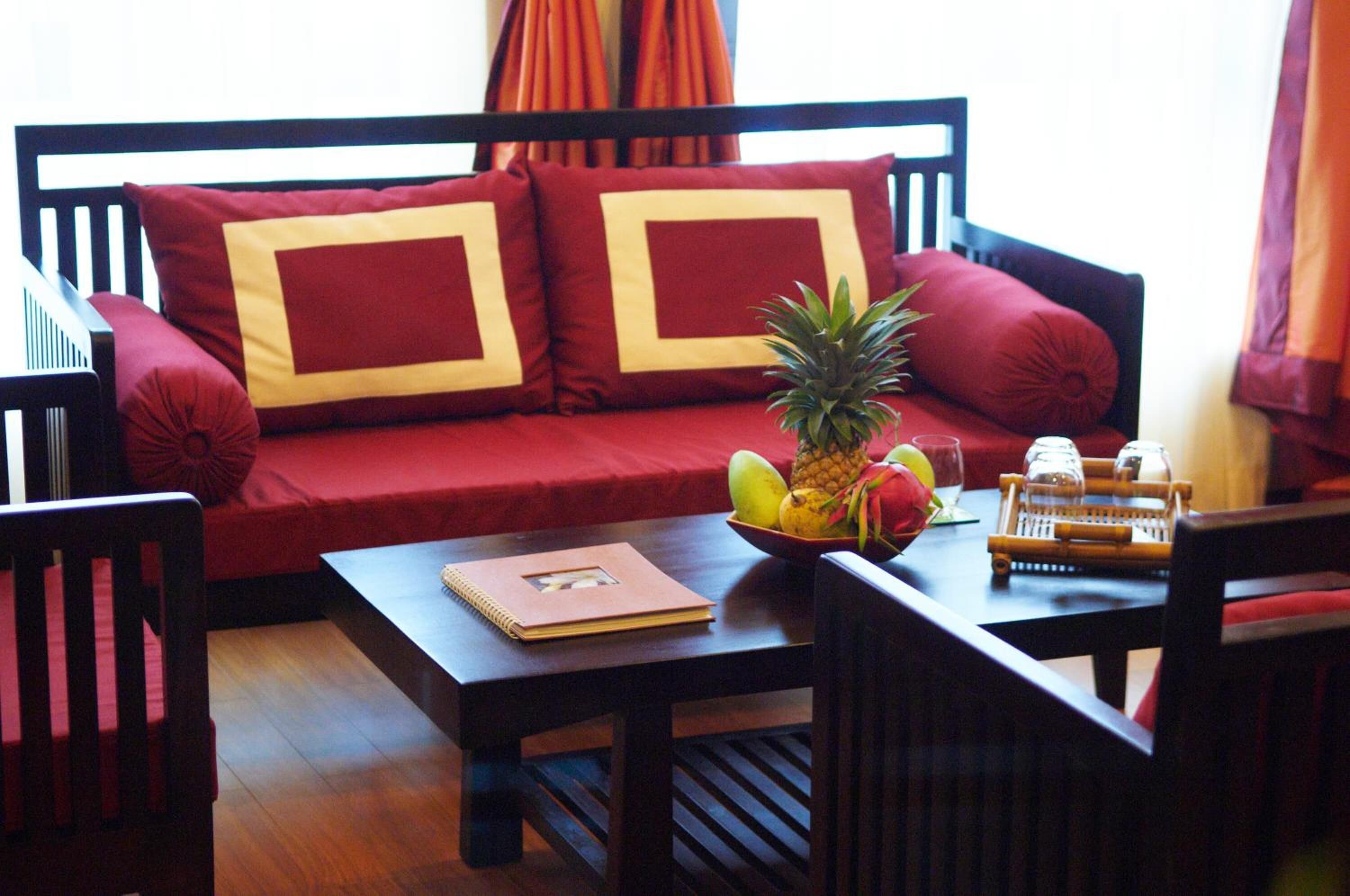 A room with a red sofa, wooden chairs, a small table with a bowl of fruit and a notebook.