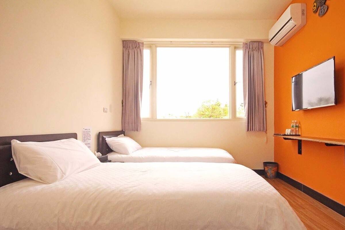 A hotel room with two beds, a TV, an air conditioner, and a window with curtains.