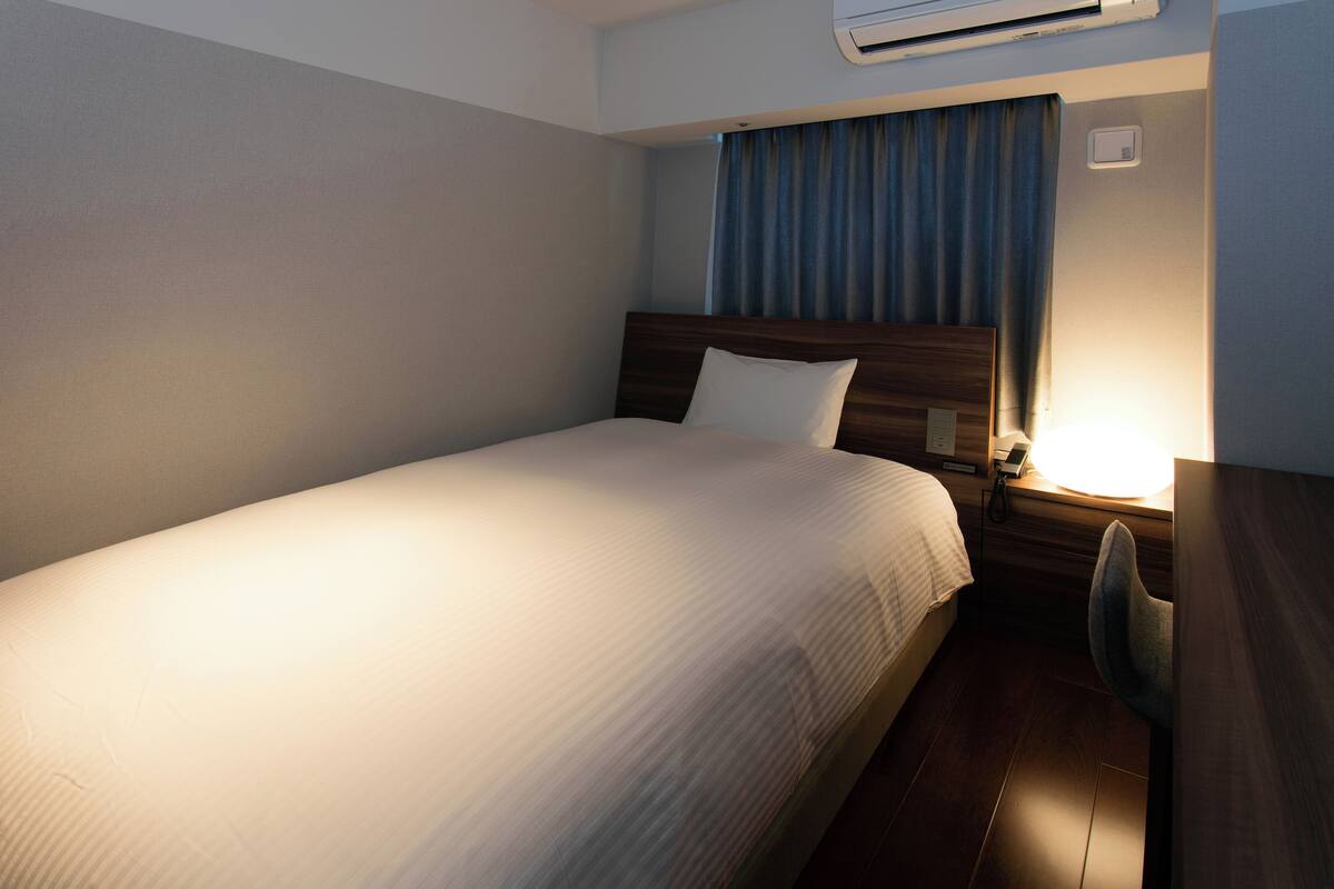 Una cama bien hecha con cabecera de madera, una mesita de noche con lámpara y un pequeño escritorio con silla.