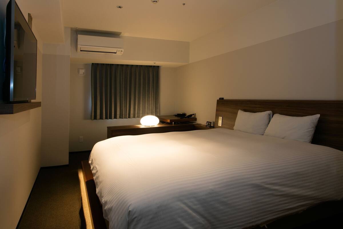 Habitación de hotel con una cama grande, un televisor de pantalla plana montado en la pared, un equipo de aire acondicionado y una mesita de noche con lámpara.