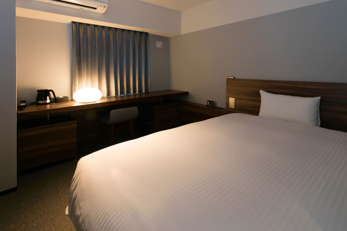 Habitación de hotel con una cama grande, un escritorio con lámpara, una silla y una ventana con cortinas.