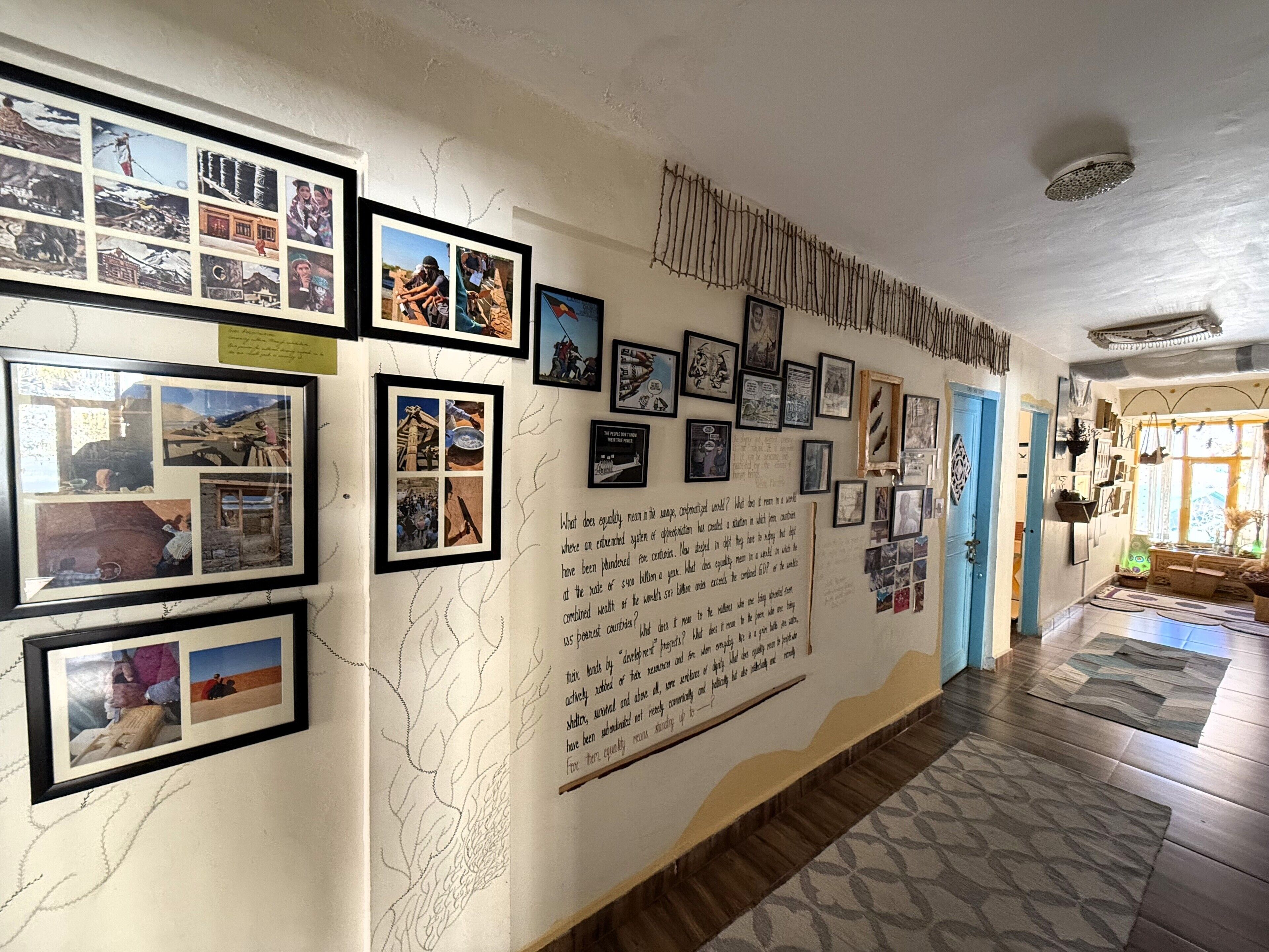 Een gang met ingelijste foto’s en tekst op de muren, die leidt naar een kamer met een blauwe deur en een raam.