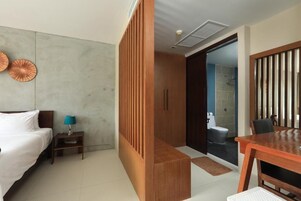 Habitación de hotel con cama, mesita de noche, lámpara, separador de madera y vistas al baño.