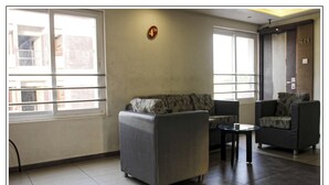 A modern living room with a sofa, two armchairs, and a small table. There are large windows with blinds, a door with a number sign, and a tiled floor. - Hotel Prem Nivas (Madurai)